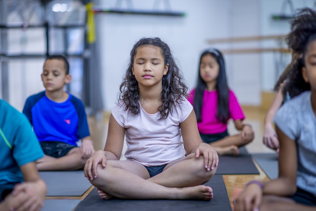A group of children sit cross-legged and meditate on mats, practicing mindfulness.