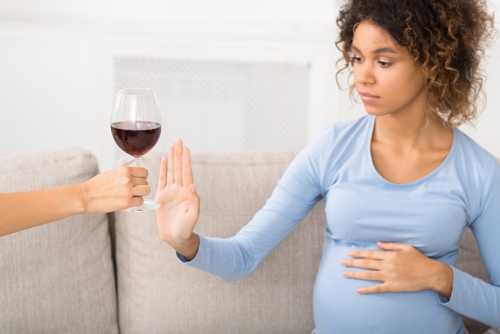 No alcohol. Pregnant woman gesturing stop to offered glass of wine, sitting on sofa.