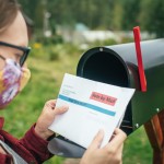 Woman in protective face mask sending or receiving letter with voting ballot near a mailbox near her house