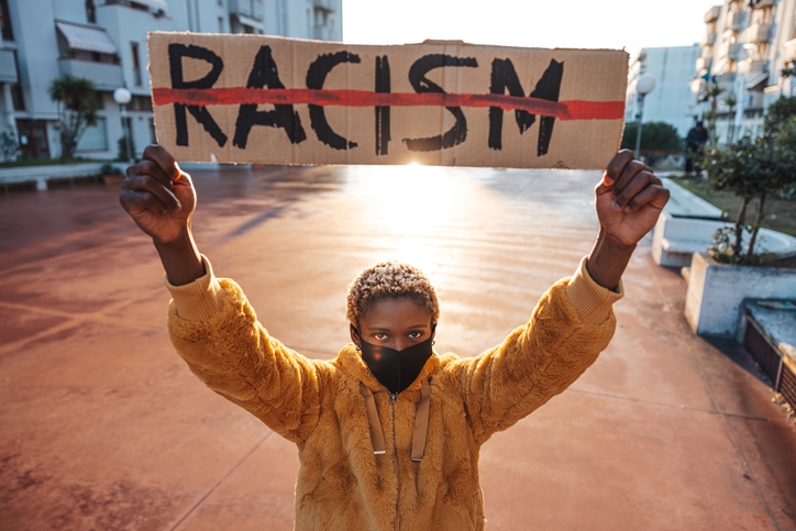 person wearing a face mask holds a placard with the word racism crossed out