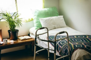 Image description: Photo of an empty bed in the corner of a room, topped by two pillows and a handmade blanket. A walker is positioned next to the bed, and sunlight is coming through a window with a curtain. The window is over a low table that holds a mug, plant, and open book.