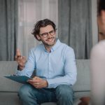 Happy european millennial guy psychologist in glasses gesturing, consulting female client in clinic