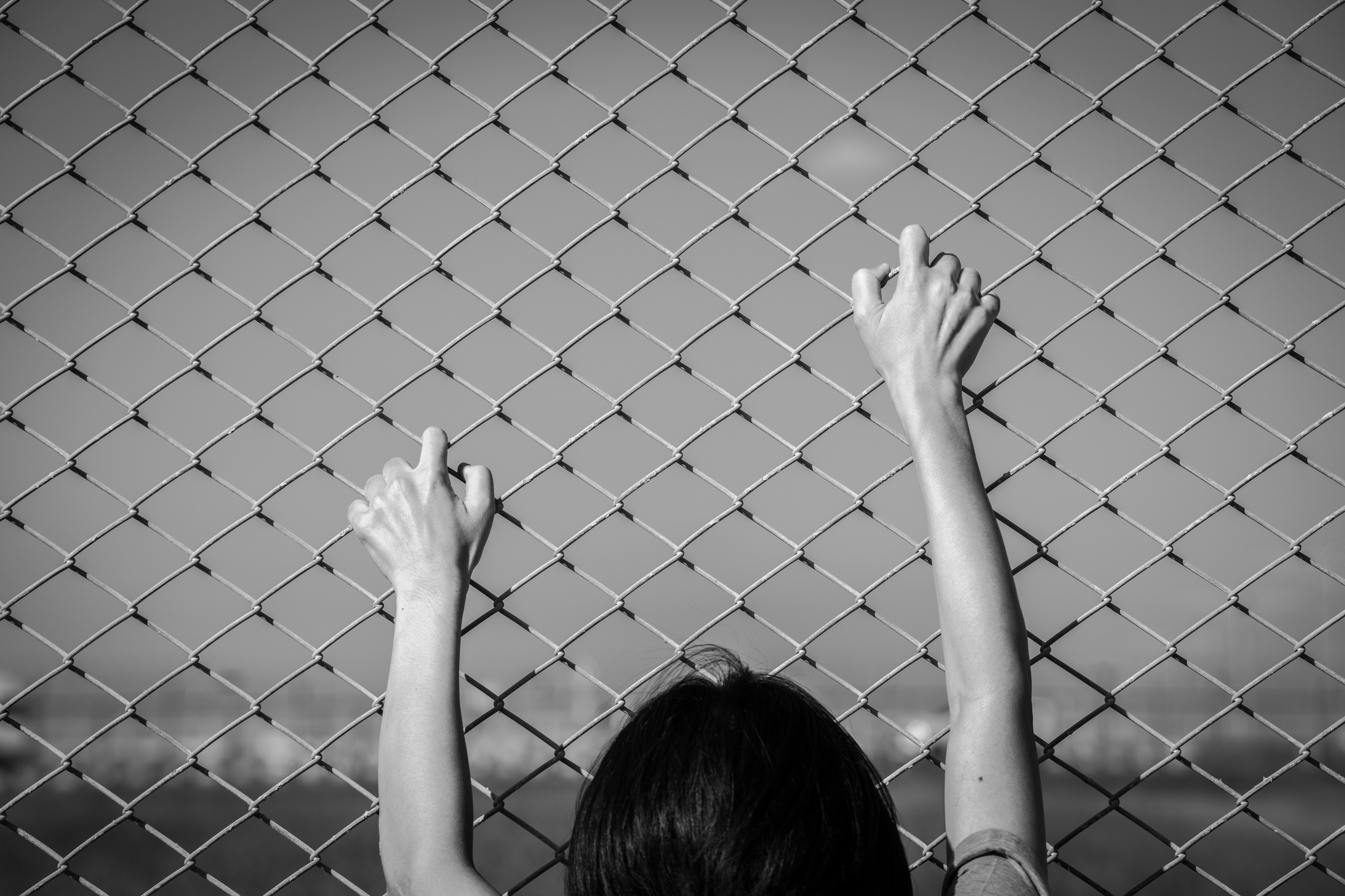 Closeup of a human hand gripping a chain link fence as if trying to escape, representing border control, imprisonment, human trafficking, and the fight against restricted movement and injustice.