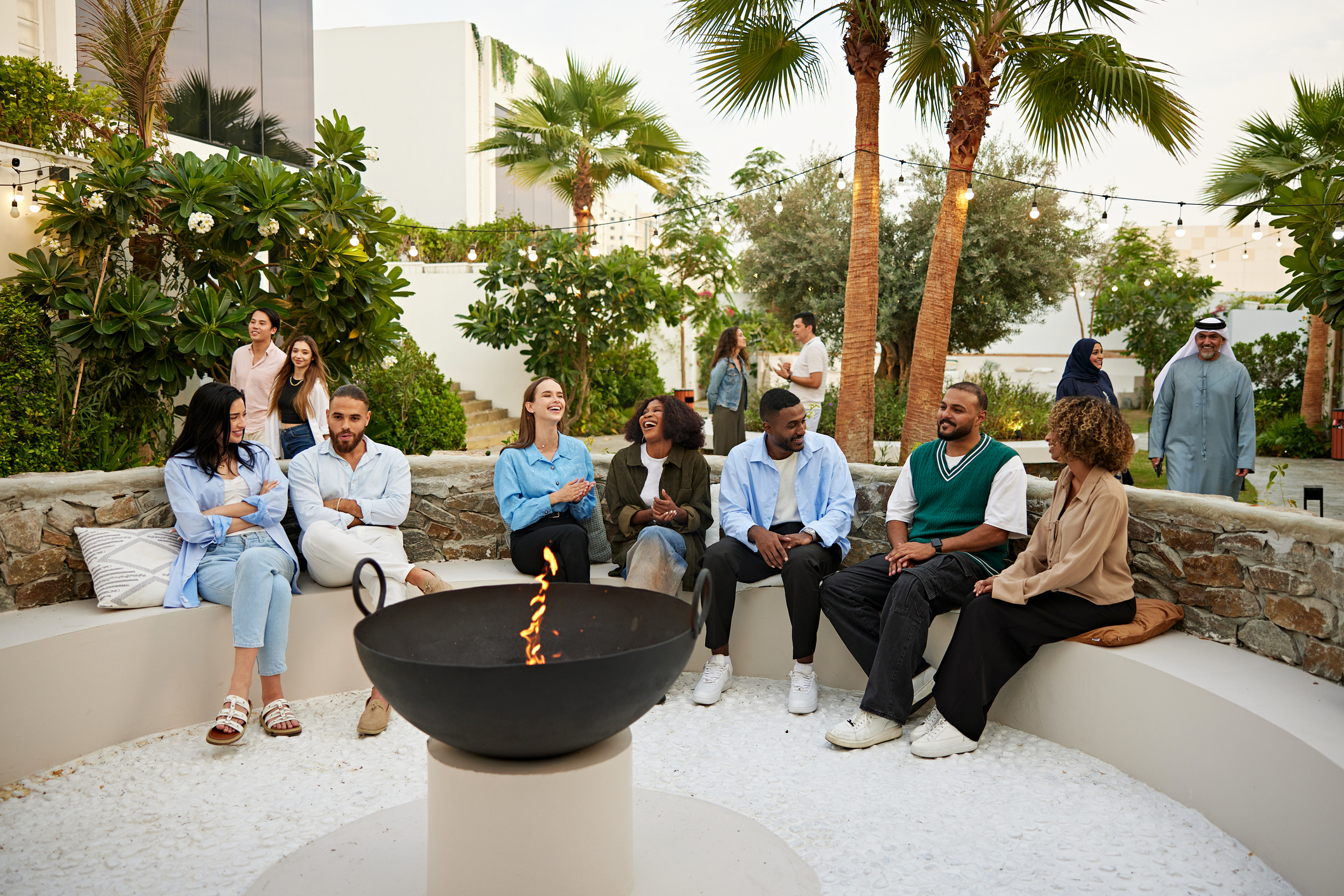 A multi racial group of employees relax around a fire pit.