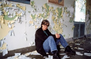 woman in jeans sits on dirty floor, leaning on wall with paint peeling and flaking off